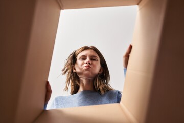Beautiful woman opening cardboard box puffing cheeks with funny face. mouth inflated with air, catching air. - obrazy, fototapety, plakaty