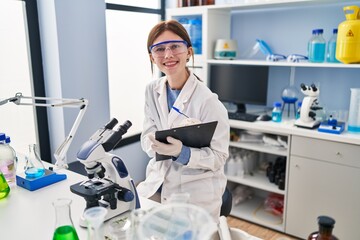 Young blonde woman scientist using microscope writing on document at laboratory