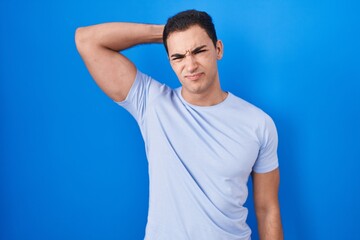 Young hispanic man standing over blue background confuse and wondering about question. uncertain with doubt, thinking with hand on head. pensive concept.