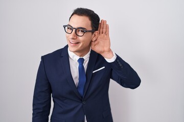 Young hispanic man wearing suit and tie smiling with hand over ear listening an hearing to rumor or gossip. deafness concept.