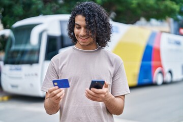 Young latin man using smartphone and credit card at bus station