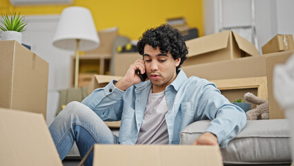 Young latin man talking on smartphone unpacking cardboard box looking upset at new home