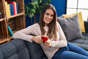 Young beautiful hispanic woman using smartphone sitting on sofa at home