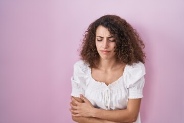 Hispanic woman with curly hair standing over pink background with hand on stomach because nausea,...