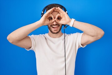 Hispanic man with beard listening to music wearing headphones doing ok gesture like binoculars sticking tongue out, eyes looking through fingers. crazy expression.