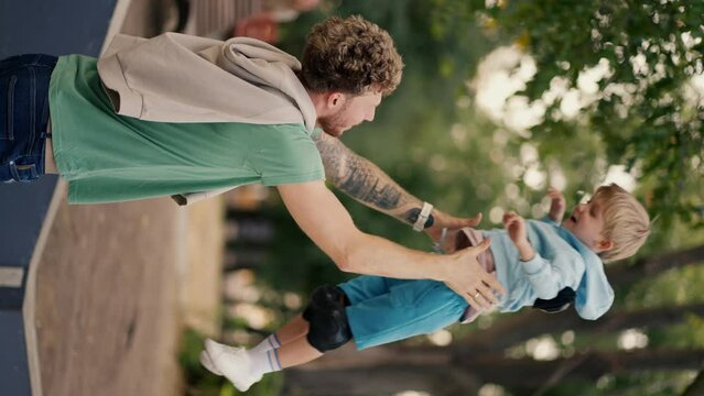 Vertical video of a dad in a Green T-shirt throwing his little blond son in blue clothes on a skate park in the summer in the park