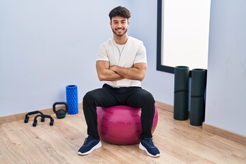 Hispanic man with beard sitting on pilate balls at yoga room happy face smiling with crossed arms looking at the camera. positive person.