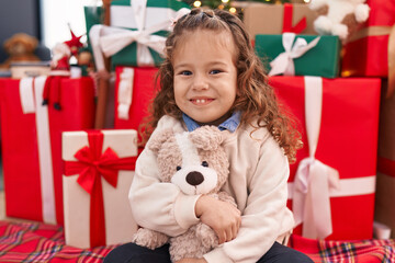 Fototapeta na wymiar Adorable blonde toddler hugging teddy bear sitting on floor by christmas tree at home