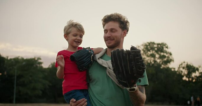 Happy little blond boy in a red T-shirt holds a baseball in his hands and throws it into the glove of his dad in a Green T-shirt. Portrait of a boy and his dad playing baseball in the park