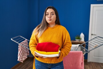 Young hispanic woman holding clean and folded laundry puffing cheeks with funny face. mouth inflated with air, catching air.
