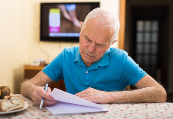 Concentrated elderly man filling up papers at home table - obrazy, fototapety, plakaty