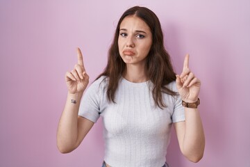 Young hispanic girl standing over pink background pointing up looking sad and upset, indicating...