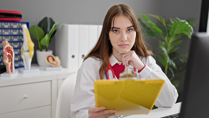 Young hispanic woman doctor reading medical report with serious expression at clinic