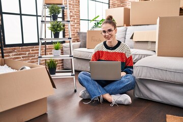 Young hispanic girl sitting on the floor at new home with laptop looking positive and happy standing and smiling with a confident smile showing teeth