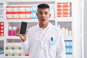 Young hispanic man working at pharmacy drugstore showing smartphone screen scared and amazed with open mouth for surprise, disbelief face