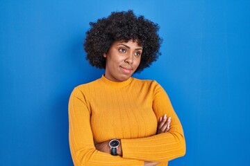 Obraz na płótnie Canvas Black woman with curly hair standing over blue background looking to the side with arms crossed convinced and confident
