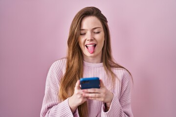 Young caucasian woman using smartphone typing message sticking tongue out happy with funny...