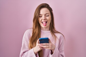 Young caucasian woman using smartphone typing message sticking tongue out happy with funny expression.