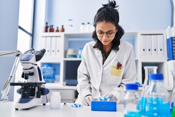 Young latin woman scientist writing on notebook holding pills at laboratory