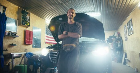 Portrait of a confident motorist with collected hair in a gray T-shirt who looks at the camera and leans on the hood of his car in his garage workshop