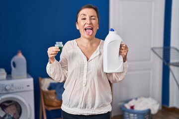 Middle age hispanic woman holding detergent bottle sticking tongue out happy with funny expression.