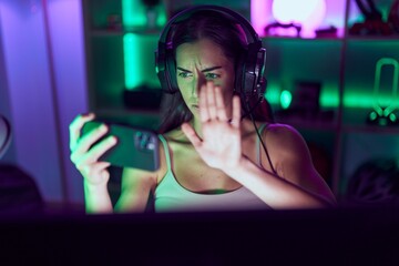 Young brunette woman playing video games with smartphone with open hand doing stop sign with serious and confident expression, defense gesture