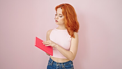 Young redhead woman holding book with serious expression over isolated pink background
