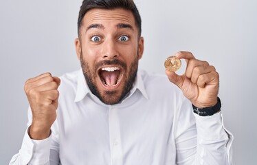 Handsome hispanic man holding monero cryptocurrency coin screaming proud, celebrating victory and success very excited with raised arms