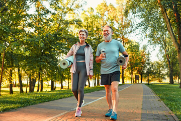 Beautiful sporty healthy active cheerful smiling middle-aged couple going to workout outdoors in park holding mats for yoga, pilates, gym. Sports healthy lifestyle. Friends on a morning walk. - obrazy, fototapety, plakaty