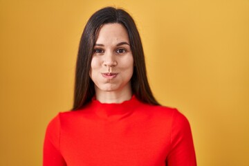 Young hispanic woman standing over yellow background puffing cheeks with funny face. mouth inflated with air, crazy expression. - obrazy, fototapety, plakaty