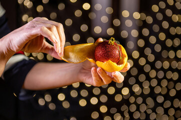 A woman from an orange peeling a Christmas ball. Closeup of hands on a background of bokeh lights.