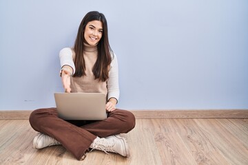 Young brunette woman working using computer laptop sitting on the floor smiling friendly offering handshake as greeting and welcoming. successful business.