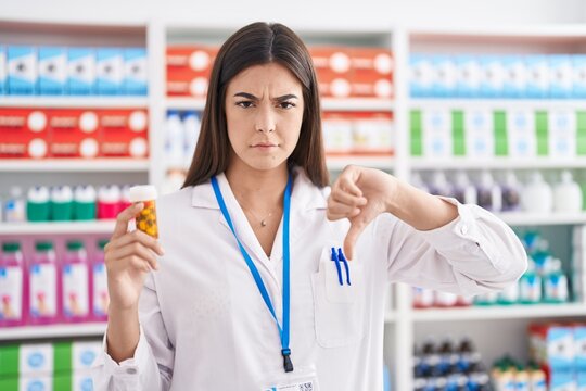 Hispanic woman working at pharmacy drugstore holding pills with angry face, negative sign showing dislike with thumbs down, rejection concept