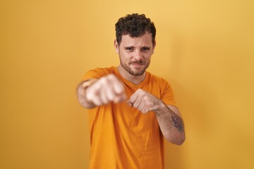 Young hispanic man standing over yellow background punching fist to fight, aggressive and angry...