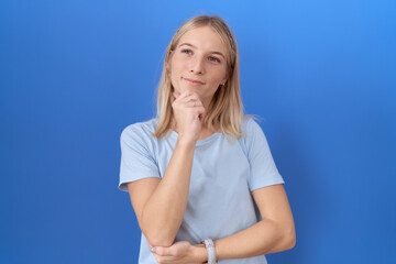 Young caucasian woman wearing casual blue t shirt looking confident at the camera smiling with crossed arms and hand raised on chin. thinking positive.