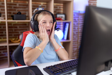 Young caucasian woman playing video games wearing headphones afraid and shocked, surprise and amazed expression with hands on face