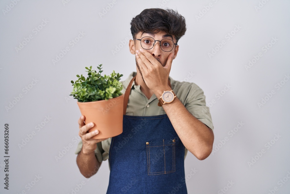 Sticker arab man with beard holding green plant pot shocked covering mouth with hands for mistake. secret co