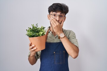 Arab man with beard holding green plant pot shocked covering mouth with hands for mistake. secret concept.