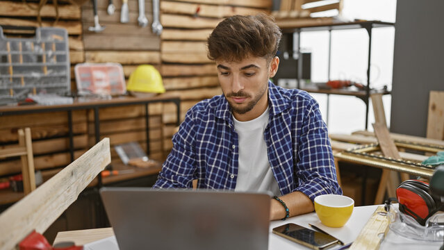 Handsome young arab man, professional carpenter, using laptop at his timber workshop. engaged in his woodworking business, masterly shaping furniture.