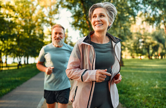 Cardio workout concept. Spirited sport couple jogging in morning in urban park during sunny day. Senior man and woman in activewear keeping in shape through regular training.