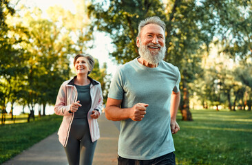 Running in park at morning time. Cheerful husband and wife competing together and jogging on fresh air. Active people wearing sport clothes doing cardio for good health and staying fit. - obrazy, fototapety, plakaty