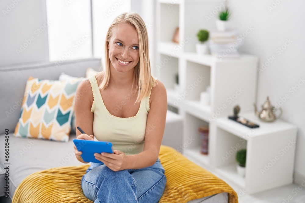 Sticker Young blonde woman using touchpad sitting on sofa at home