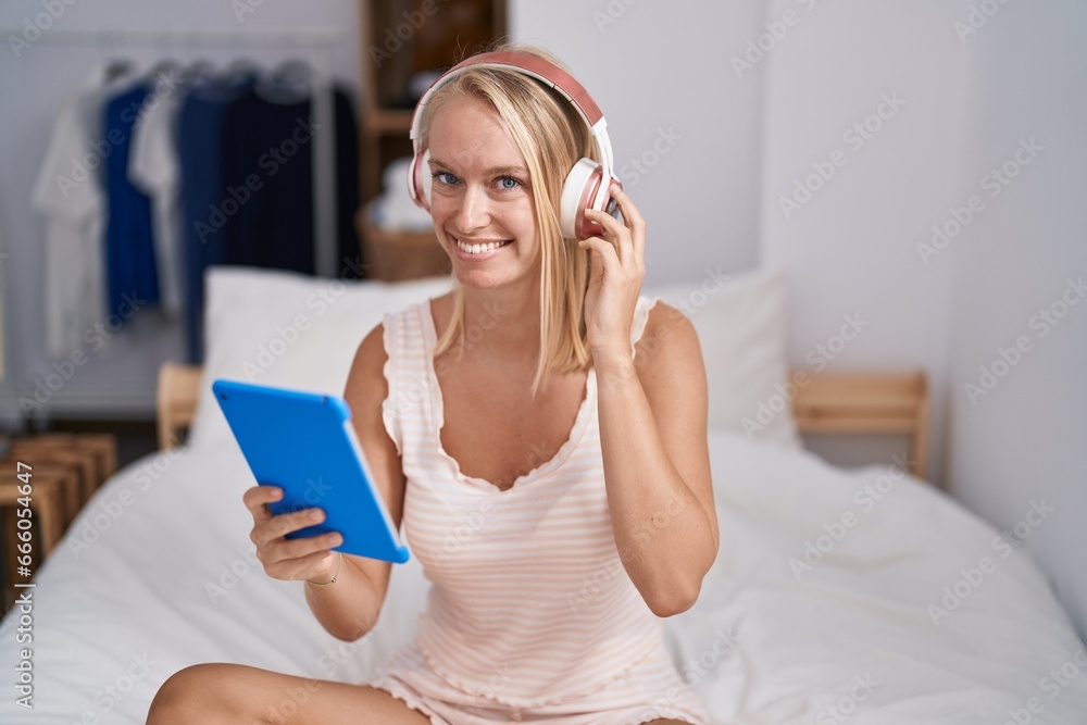 Poster Young blonde woman using touchpad and headphones sitting on bed at bedroom