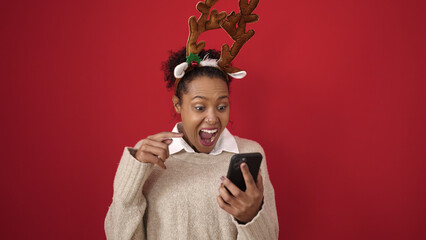 African american woman using smartphone wearing reindeer ears over isolated red background