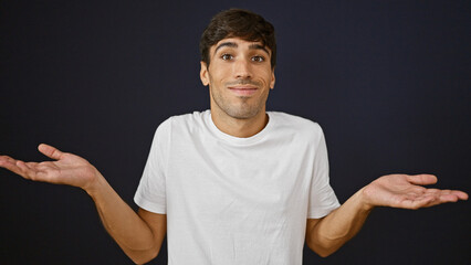 Confused young hispanic man cluelessly standing alone against isolated black background, this...