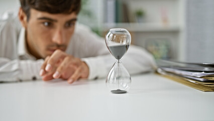 Young hispanic man business worker looking at hourglass at the office