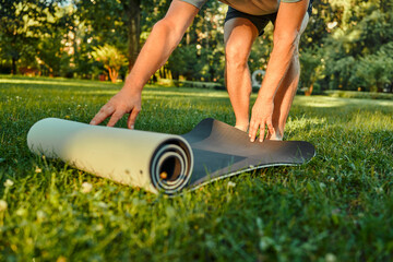 Exercising on fresh air. Crop of male person standing on freshly cut lawn and spreading out black fitness rug. Athletic man getting ready for outdoor workout during sunny day. - obrazy, fototapety, plakaty