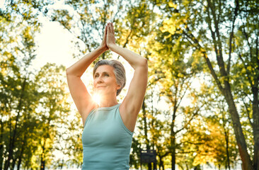 Yoga practice outdoors. Graceful aged lady keeping hands clasped above head and making regular workout on fresh air. Grey-haired woman wearing sport attire performing vrksasana tree pose.