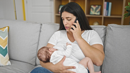 Mother and baby daughter sitting on sofa breastfeeding speaking on the phone at home