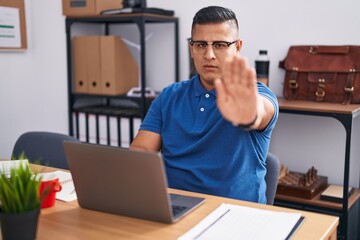 Young hispanic man working at the office with laptop doing stop sing with palm of the hand. warning expression with negative and serious gesture on the face.